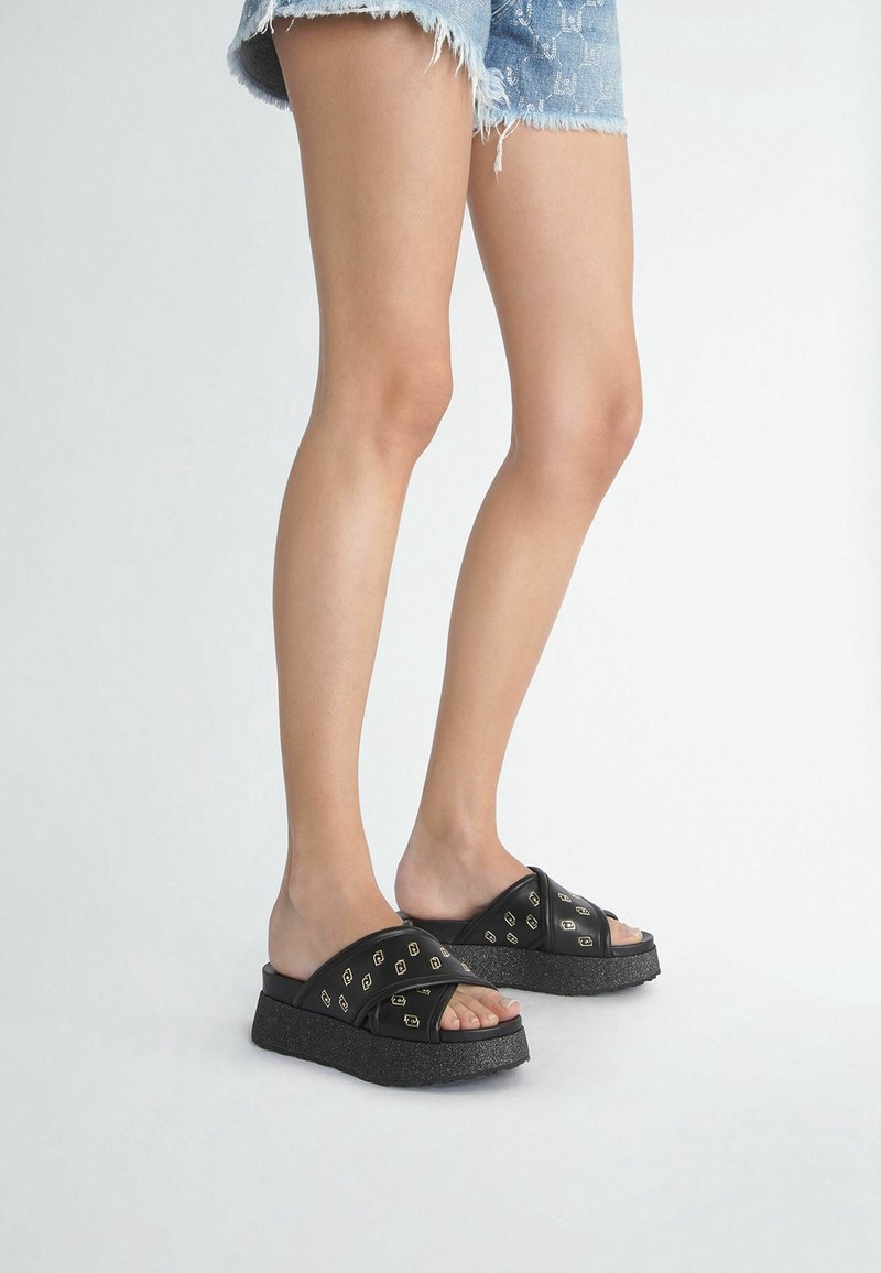 Piernas con pantalones cortos de mezclilla desgastados y sandalias de plataforma negras con correas de patrón de cadena dorada sobre un fondo blanco liso.