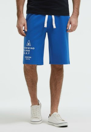Mann trägt blaue Shorts mit Kordelzug, weißem Text und Logo am linken Bein, weiße Sneakers und ein schwarzes Shirt, steht vor weißem Hintergrund.