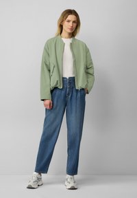 Cazadora bomber verde claro con cierre de cremallera, combinada con vaqueros ajustados de cintura alta en azul. El modelo lleva una camiseta blanca y zapatillas grises.