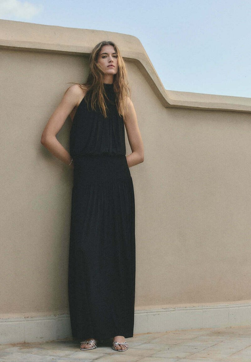 Jeune femme aux longs cheveux portant une robe maxi noire sans manches et des sandales argentées, debout contre un mur beige sous un ciel dégagé.