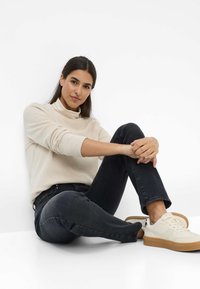 Maglione beige a coste con collo alto abbinato a jeans scuri e sneaker crema. Modella seduta con le gambe incrociate su uno sfondo bianco.
