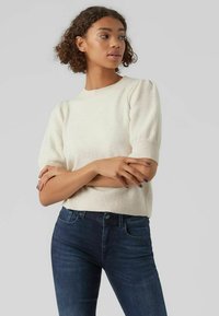 Krämfärgad stickad tröja med korta puffärmar och rund halsringning, kombinerad med mörkblå denimjeans. Mjuk textur och avslappnad passform.
