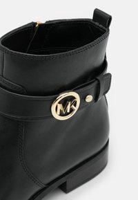 Bottine en cuir noir avec un logo MK doré sur une bride, une texture lisse et un talon bloc bas. Fermeture éclair à l'arrière.
