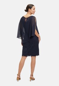 Námorně modré šaty s krajkovou sukní a průhledným pláštěm. Mají vykrojený zadní díl a zvlněný lem. Boty jsou tanové sandály na podpatku.