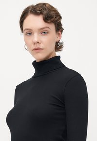 Young person with short wavy hair and hoop earrings wearing a black ribbed turtleneck against a plain white background.