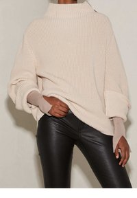 Pull en maille beige avec un col haut, une texture côtelée et des manches retroussées, associé à un pantalon noir ajusté et brillant.