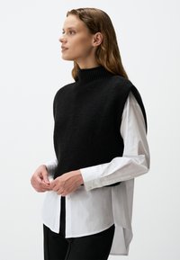 Zwarte gebreide mouwloze trui met hoge kraag, gedragen over een witte blouse met knopen. De top heeft een gestructureerde afwerking en een losse pasvorm.
