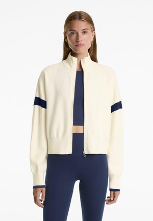 Cremefarbener Reißverschluss-Pullover, gerippte Bündchen, marineblaue Akzentstreifen an den Ärmeln, weiche Strickqualität, lockere Passform, getragen über einem marineblauen Oberteil.