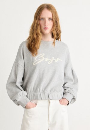 Felpa grigia con "Boss" ricamato in crema. Presenta un collo rotondo, maniche arricciate e un cinturino aderente in vita. Indossata con pantaloni bianchi.