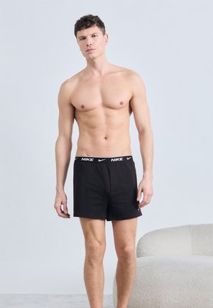 Ing nélkül álló férfi, fekete Nike logós derékpántos rövidnadrágban, világos bőrű, rövid göndör hajjal, semleges arckifejezéssel, jobb oldalon fehér ívelt kanapé.