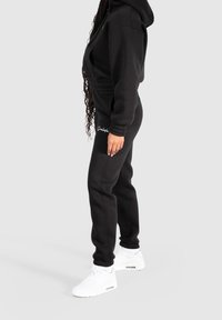 Black hoodie and pants set made of soft fabric, featuring ribbed cuffs and waistband. Accented with white logo detail on the pants.