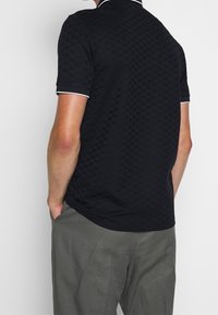 Man med en svart texturerad polo skjorta med vita detaljer vid ärmar och krage, i kombination med lösa grå byxor, med händerna i fickorna, vänd bort.