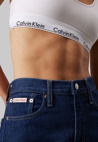 Jeans de denim de Calvin Klein con un lavado azul oscuro, que presentan un parche con el logo, herrajes plateados y un detalle de costura en contraste. Top corto de algodón blanco debajo.