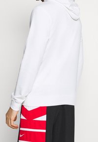 Vit hoodie med huva, med en mjuk textur. Bärs med svarta shorts som har ett röd- och vit geometriskt mönster.