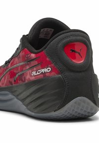 Chaussure de sport noire et rouge avec un dessus texturé, dotée d'une semelle en caoutchouc lisse, logo Puma et motif camouflage sur les côtés.