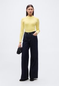 Κίτρινη μακρυμάνικη μπλούζα από πλεκτό ύφασμα με ριγέ υφή, συνδυασμένη με σκούρο μπλε τζιν παντελόνι φαρδύ στο πόδι, τονισμένη με μαύρη ζώνη και μικρή μαύρη τσάντα χειρός.