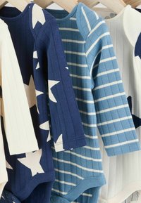 Trois bodies pour bébé accrochés sur des cintres en bois : un crème avec des étoiles, un à côtes bleu foncé et un rayé bleu clair. Texture en coton doux.