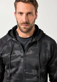 Zwarte camouflage zip-up hoodie met een trekkoordcapuchon, gestructureerde stof en een aangesloten ontwerp, gelaagd over een zwart shirt.