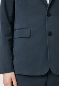 Mörkgrå kavaj i strukturerad tyg. Har en bröstficka och två svarta knappar fram.