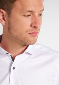 Witte shirt met een puntige kraag en zwarte knoopdetails. Beschikt over een gepersonaliseerde binnenkraag en een gladde stoftextuur.