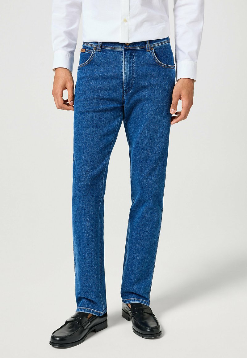 Férfi kék egyenes szárú farmernadrágban, fekete bőrbakancsban és fehér gombos ingben, egyszínű háttér előtt.