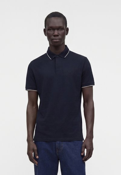 Homme portant un polo bleu marine foncé avec des bordures blanches au col et aux manches, associé à un jean bleu foncé, debout devant un fond blanc uni.