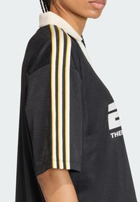 Μαύρο πόλο μπλουζάκι με λευκό γιακά, με κίτρινες και λευκές ρίγες στα μανίκια και μεγάλο λευκό λογότυπο στο μπροστινό μέρος. Μαλακό ύφασμα.