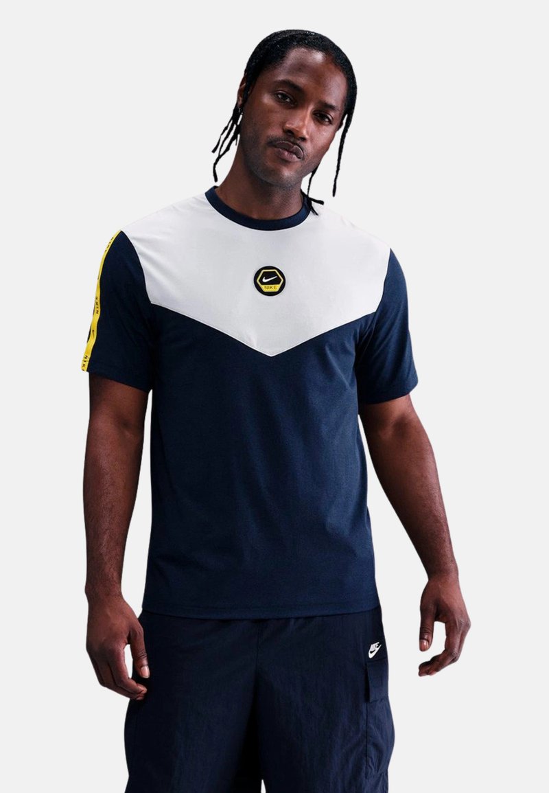 Camiseta de Nike en color azul marino y blanco con un diseño en bloque en forma de V, un parche de logo negro y amarillo en el pecho y mangas cortas.