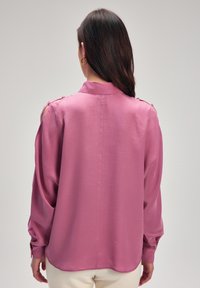 Blusa rosa a maniche lunghe con una texture morbida, cut-out sulle spalle e dettagli con bottoni. Presenta un colletto e una vestibilità rilassata.