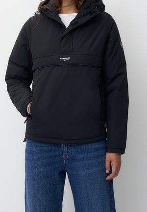 Persona con chaqueta acolchada negra con capucha y bolsillo frontal, jeans azules, de pie frente a un fondo blanco liso.