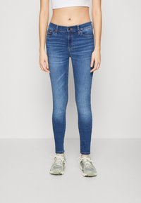 Pantalones vaqueros ajustados de mezclilla azul con cintura de altura media, un ligero desvanecimiento y diseño de cinco bolsillos. Combinados con zapatillas deportivas multicolores.
