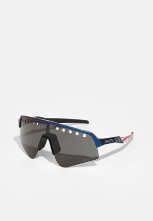 Occhiali da sole con lenti scure di grandi dimensioni, montatura in metallo blu, fori di ventilazione sulla parte superiore e asta decorata con un motivo rosso, bianco e blu.