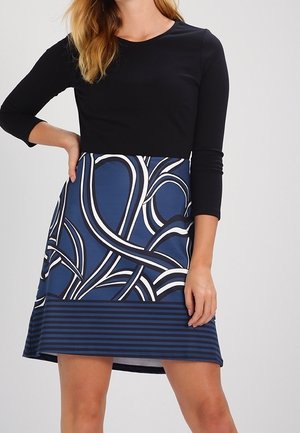 Frau trägt ein schwarzes Langarmshirt und einen blauen Rock mit abstrakten weißen und schwarzen Wirbelmustern sowie einem gestreiften Saum.