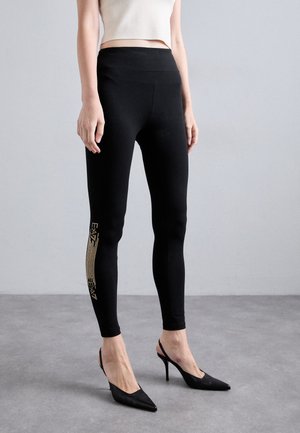 Zwarte legging gemaakt van rekbaar materiaal, met een hoge taille en sprankelende gouden details langs de zijkant. Gecombineerd met puntige zwarte hakken.