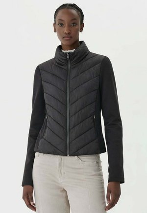 Donna che indossa una giacca trapuntata nera aderente con cerniera frontale e pantaloni beige chiaro, in piedi contro uno sfondo bianco semplice.