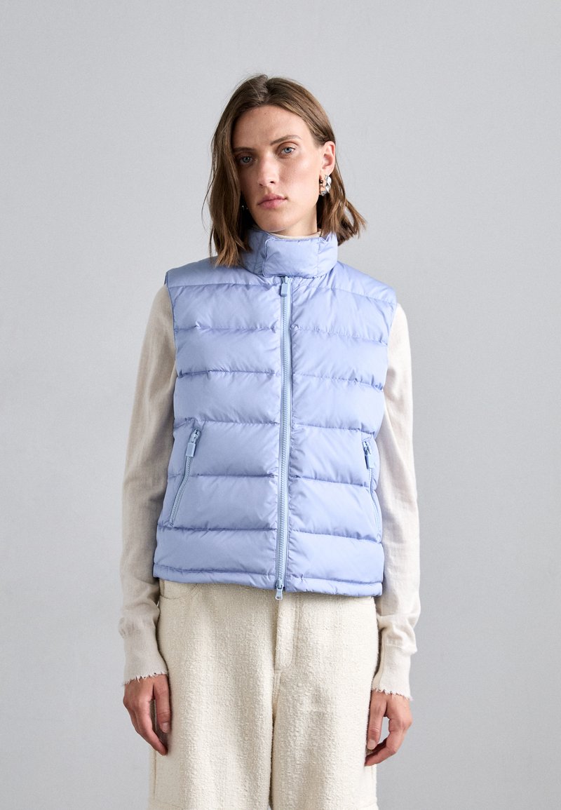 Hellblaues Pufferwesten mit hohem Kragen, horizontaler Steppung und zwei seitlichen Reißverschlusstaschen. Getragen über einem cremefarbenen Oberteil.