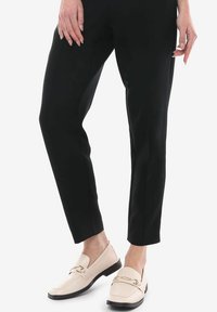 Mocasines de color crema con un acabado de cuero texturizado, que cuentan con un acento de cadena dorada y una suela negra baja, combinados con pantalones negros ajustados.