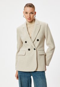 Blazer beige doppiopetto, realizzato in tessuto testurizzato, con bottoni neri, rever a punta e due tasche frontali.