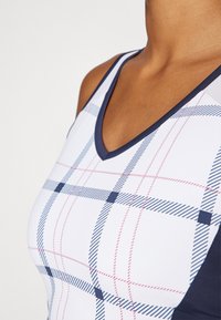 Hvit V-hals tank topp med et rutete mønster som har marineblå, rød og lys blå linjer. Glatt stoff med en tettsittende design.