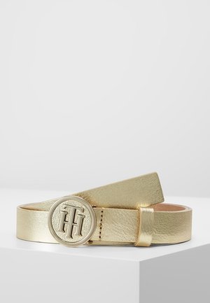 Ceinture en cuir métallique doré enroulée avec une boucle ronde dorée ornée des initiales « TH » en relief superposées, exposée sur une surface blanche.
