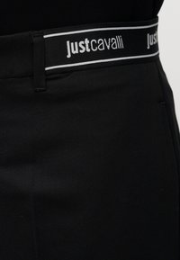 Gros plan d'un pantalon noir avec une ceinture élastique noire et blanche portant le nom de la marque « Just Cavalli » en lettres blanches.