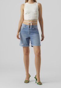 Un crop top blanc à côtes, un short en denim bleu taille haute avec des revers roulés et des accents déchirés, associé à des sandales à talons ouvertes vertes.