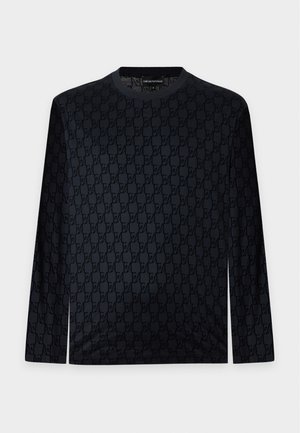 Homme portant une chemise sombre à motifs, un jean clair à jambes larges, des baskets blanches et des lunettes de soleil, debout les mains dans les poches.