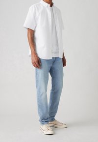 Camicia bianca a maniche corte con bottoni sopra una t-shirt bianca, abbinata a jeans celesti e sneaker beige con dettagli rossi.