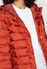 Person som har på sig en öppen, glänsande rost-orange puffjacka över en enkel vit T-shirt, som visar detaljer som tryckknappar och dragkedja på jackan.