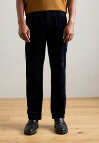 Mornarske korduroy hlače ravnog kroja, s vertikalnom rebrastom teksturom, bočnim džepovima, a nose se s crnim slip-on cipelama.