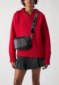 Maglione rosso a maglia con ampi polsini a costine, abbinato a una borsa a tracolla rettangolare nera con logo e tracolla regolabile. Gonna a quadri grigia.