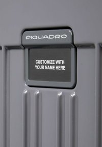 Grauer Koffer mit vertikal gerippter Oberfläche. Enthält eine anpassbare Namensplatte mit dem Text "PASSE MIT IHREM NAMEN AN".