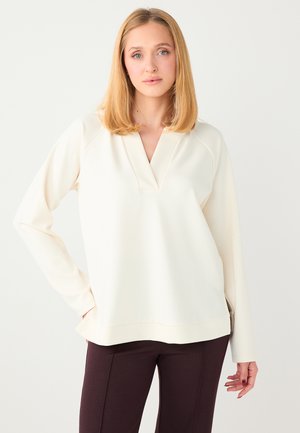 Haut à manches longues couleur crème avec un décolleté en V profond, doté de fentes latérales et d'une coupe légèrement ample, associé à un pantalon marron foncé.