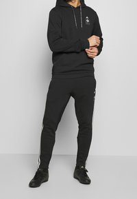 Svart hoodie med framficka och snörning; svarta sweatpants med vita sidostråk; modell som bär svarta träningsskor.